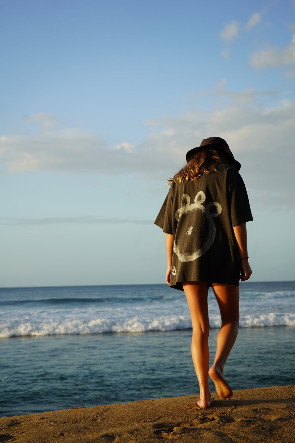 A woman stands on a sandy beach facing the ocean, wearing an oversized black graphic t-shirt with a white bear face design on the back, a dark wide-brim hat, and bare feet.