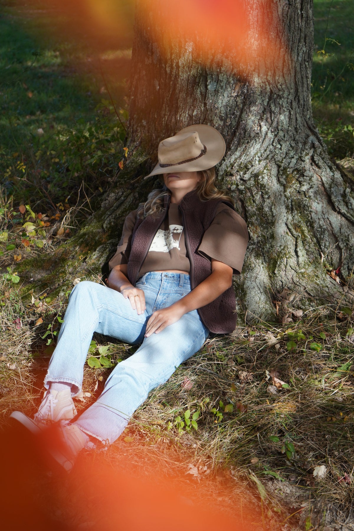 A woman sits on the ground leaning against a large tree trunk outdoors, wearing a tan cowboy hat, brown vest, brown short-sleeve top, light blue jeans, and white boots.