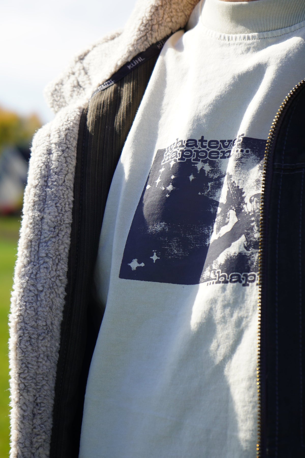 A close-up outdoor shot of a person wearing a white t-shirt with a dark blue graphic print, layered under a black zip-up jacket with a gray sherpa lining.