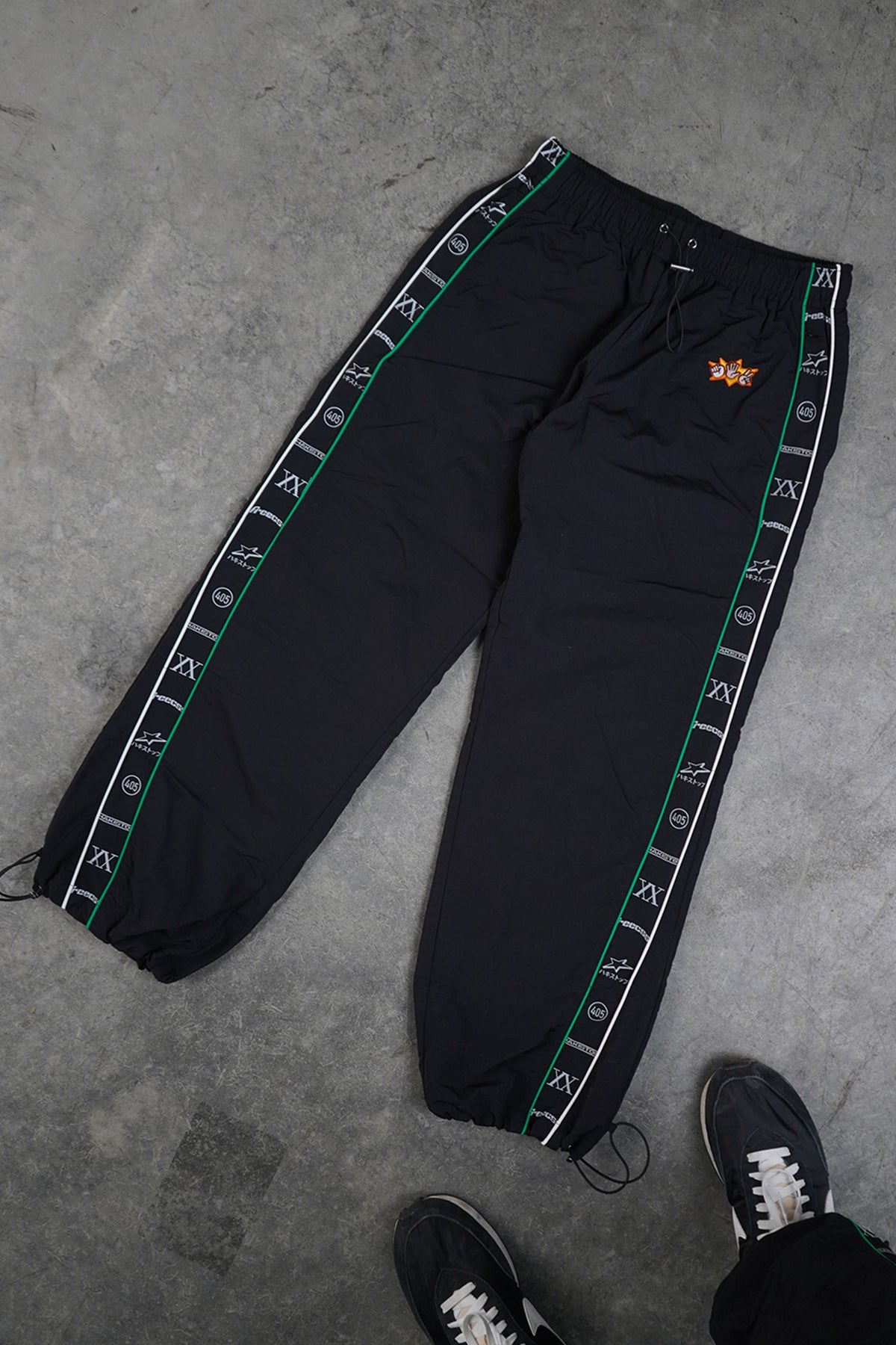 Flat lay of black Jan-ken Trackpants with green and white piped side stripes, repeating printed tape text and symbols, small orange embroidered hand-gesture patch, drawstring ankles