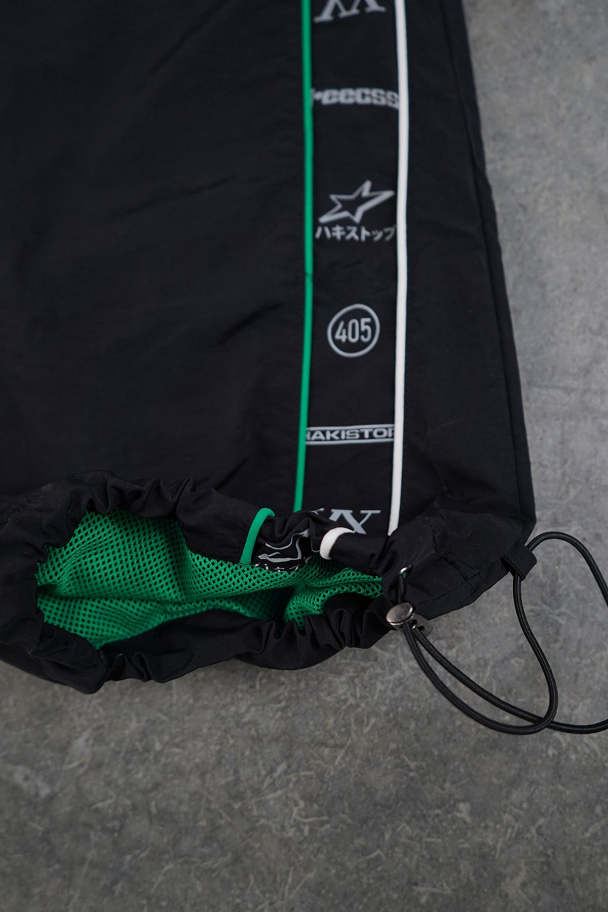Close-up of the Jan-ken Trackpants black drawstring hem with green and white piping, green mesh lining and a vertical side strip of printed text and logos