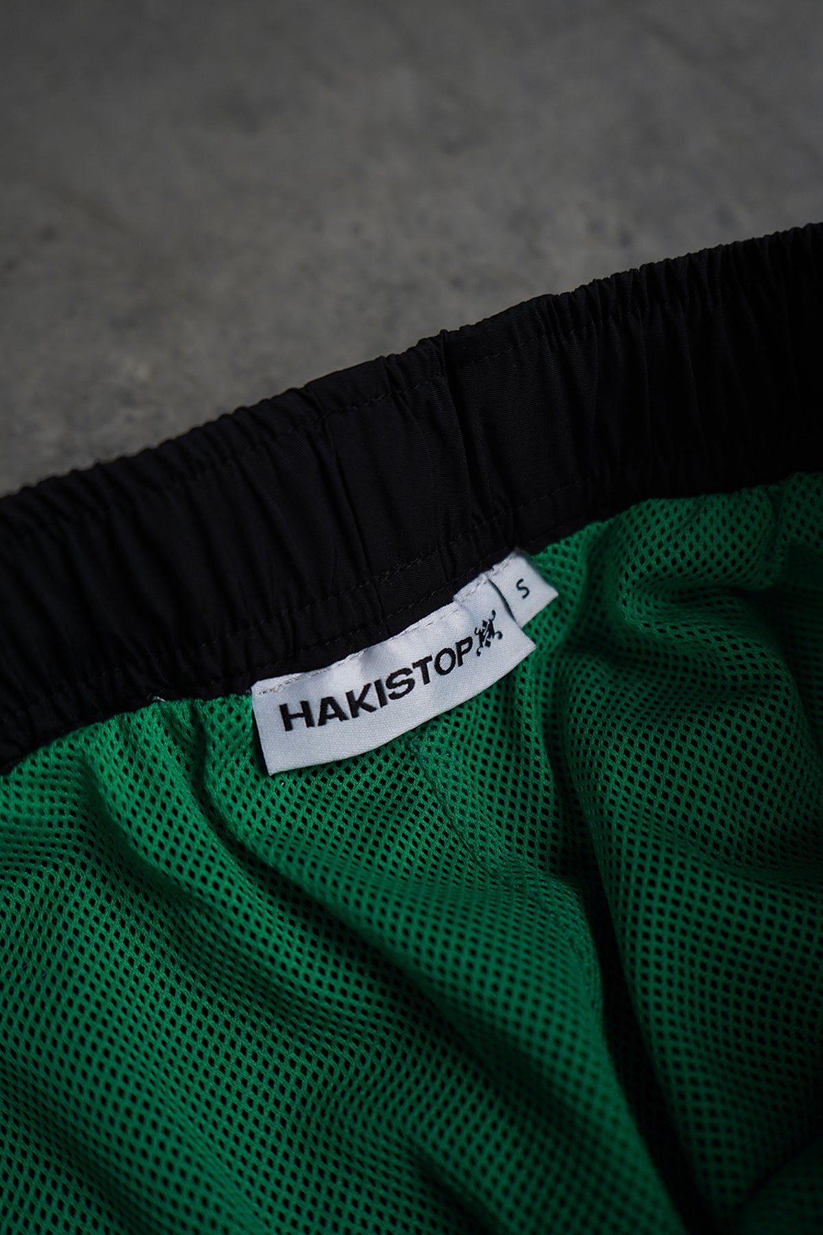 Close-up of the Jan-ken Trackpants interior: black elastic waistband, green mesh lining and a white woven HAKISTOP label in size S