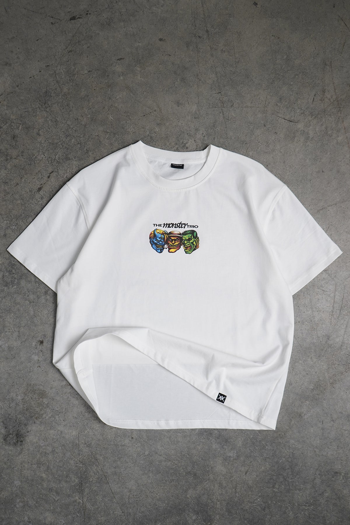 Flat lay front view of the white Monster Trio Tee with a small chest graphic of three colorful monster heads under the printed text "The Monster Trio