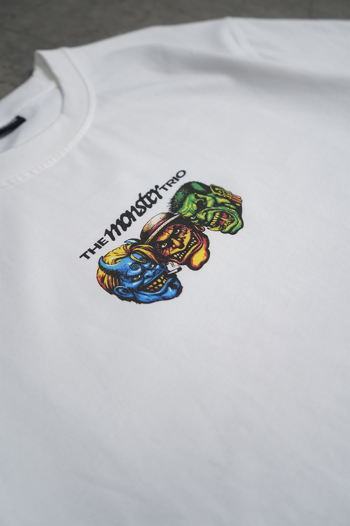 Close-up of the white Monster Trio Tee chest, showing black printed text reading "The Monster Trio" beside a graphic of three stacked colorful monster heads