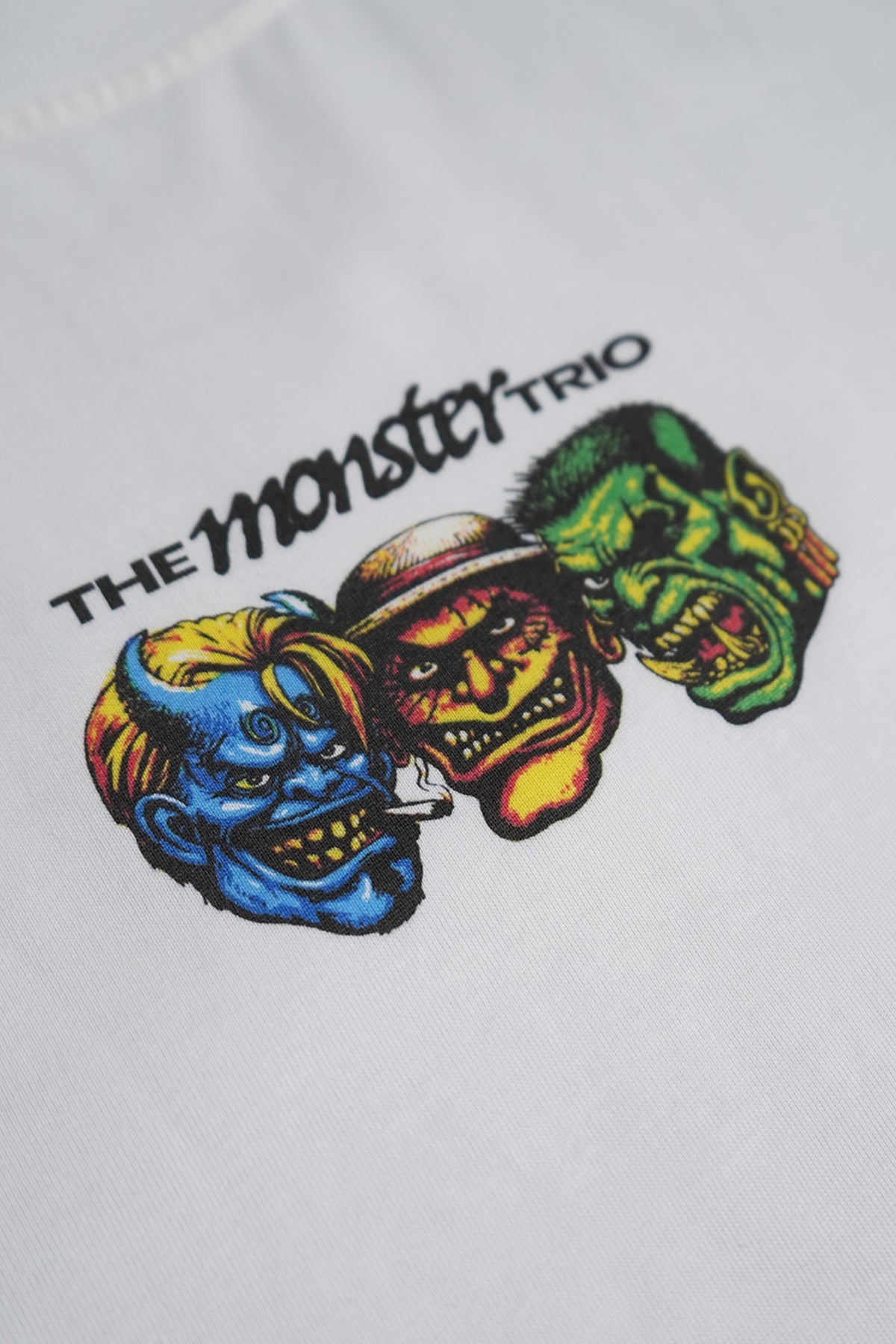Close-up of the white Monster Trio Tee showing the printed text "The Monster Trio" above three colorful grotesque monster heads: blue, red and green