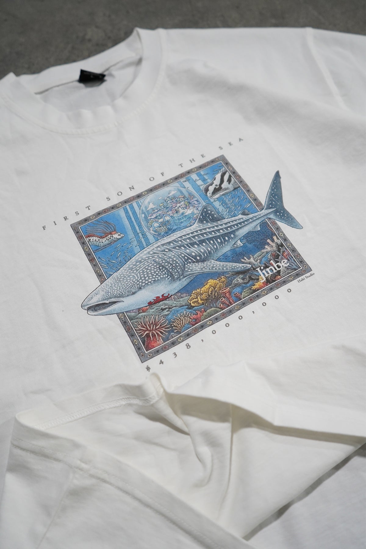 A white short-sleeve graphic T-shirt laid flat on a gray surface, featuring a large rectangular underwater print with a shark, coral reef, and small text including 'Shark'.