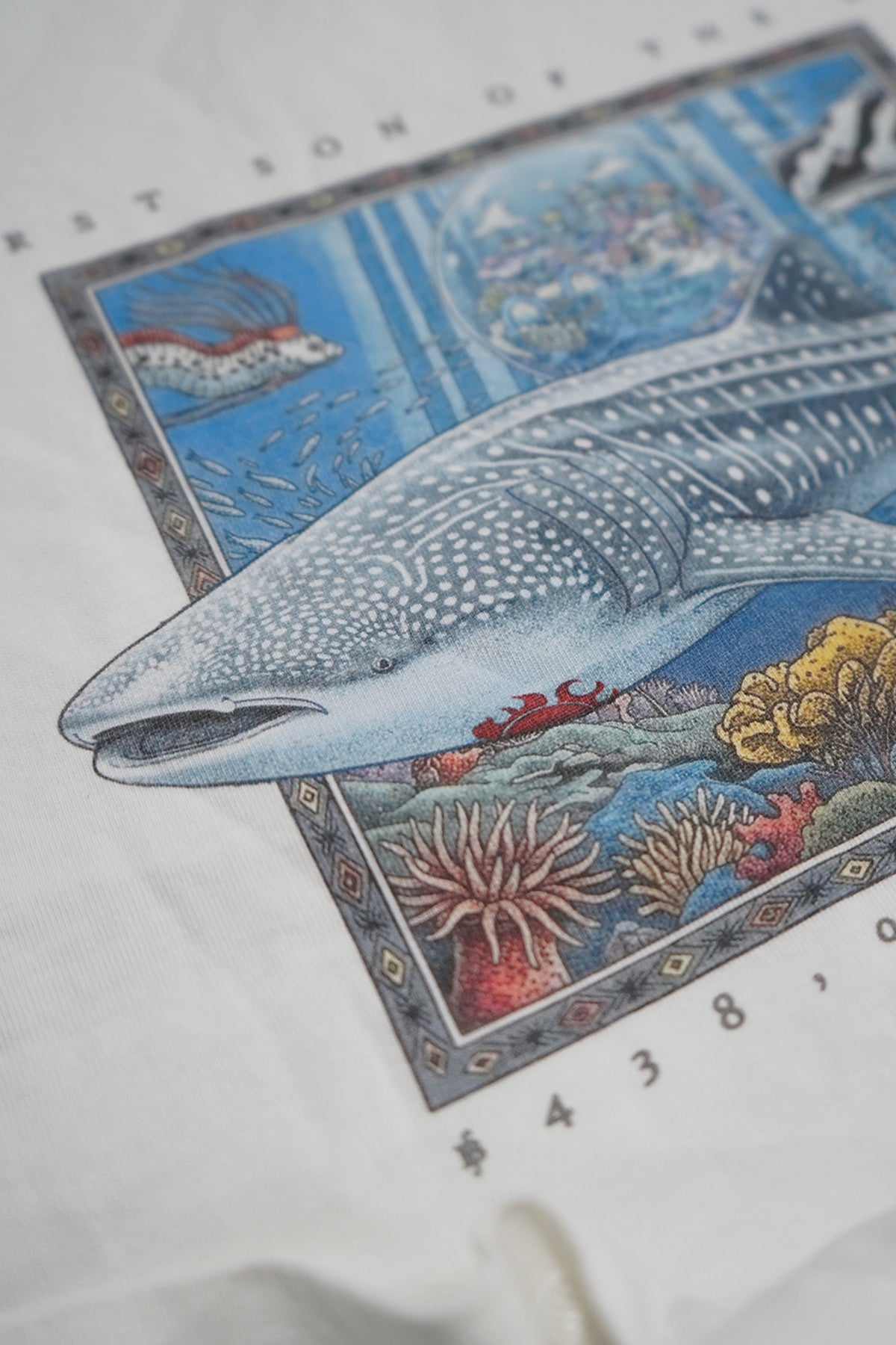 A close-up of a white fabric item, likely a t-shirt or poster print, featuring a detailed illustration of a whale shark swimming over colorful coral reefs in a blue ocean scene.