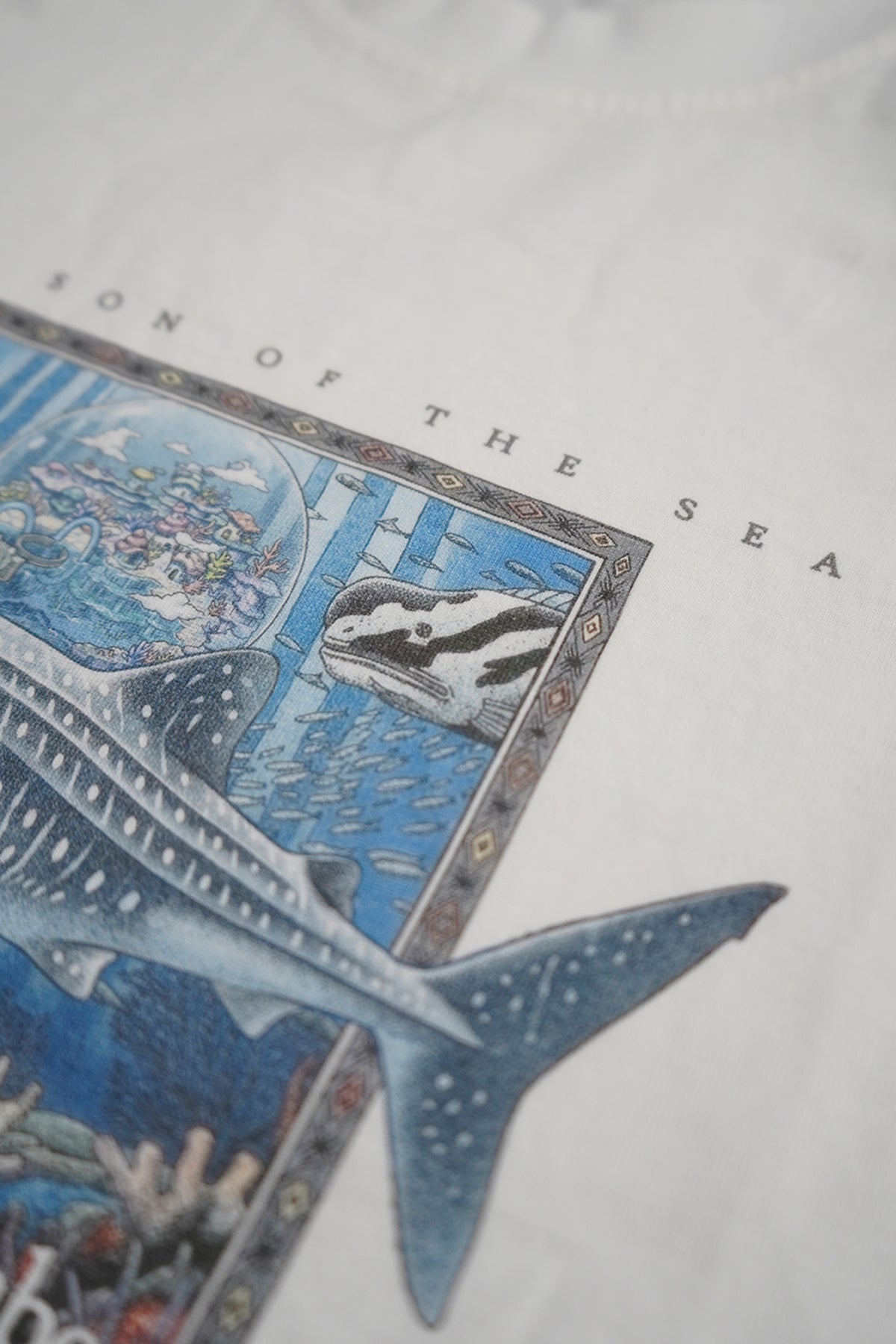 Close-up of a white T-shirt featuring a detailed graphic print of a whale shark and a sea otter in blue ocean tones, with partial text reading 'SON OF THE SEA'.