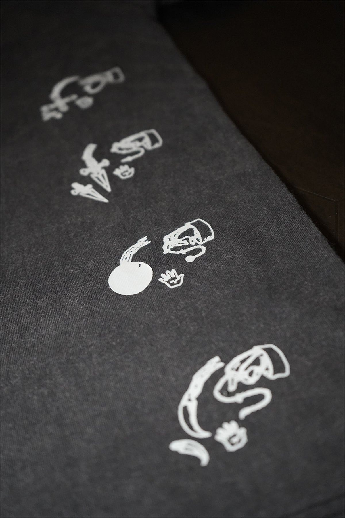Close-up of the charcoal grey Chain User Tee showing a diagonal row of small white line-art graphics, including daggers, a ball on a chain, and hands