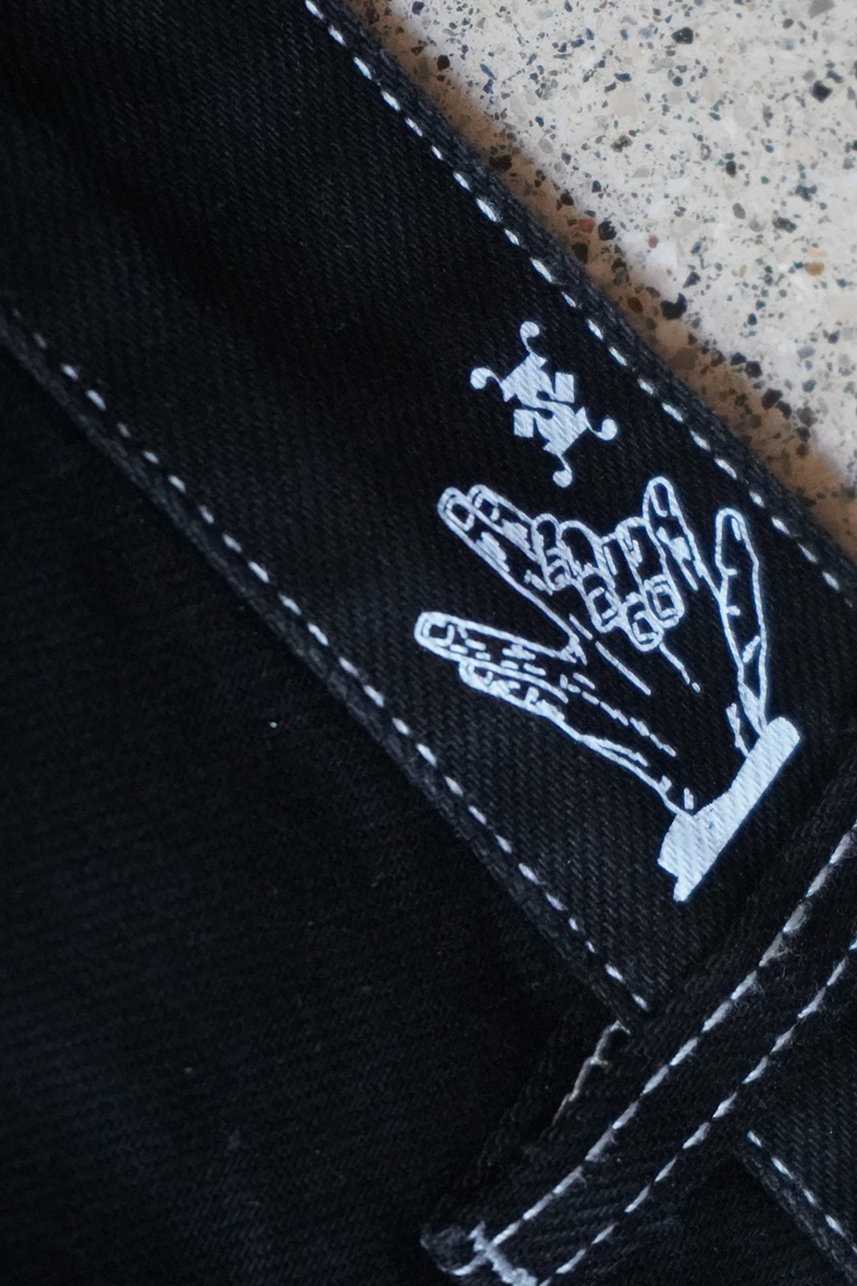 Close-up of the black Divine Dogs Pants showing white contrast stitching and a white printed graphic of a hand with spread fingers above a small ornamental symbol