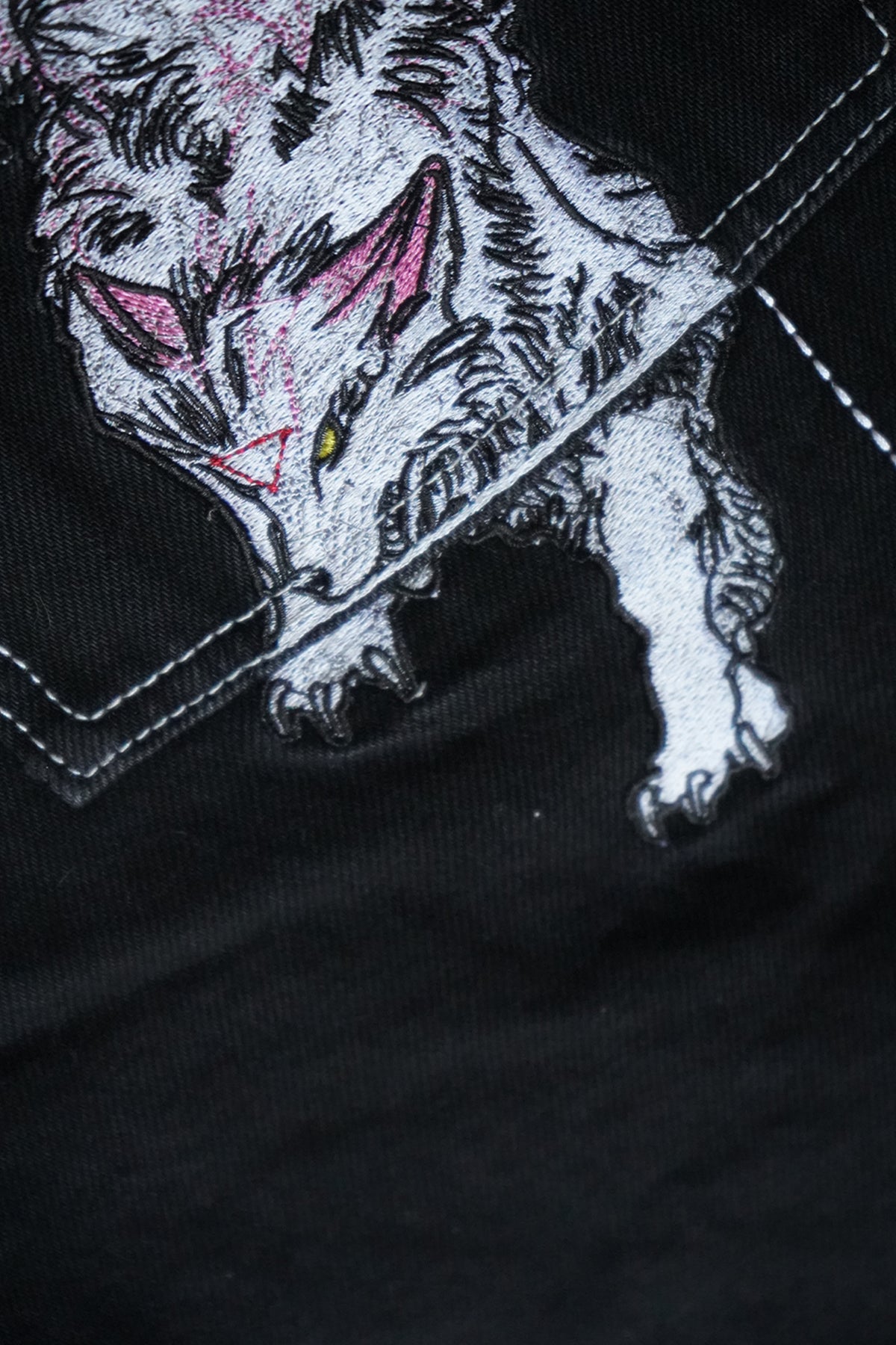 Close-up of the black Divine Dogs Pants showing an embroidered white wolf with pink ears and yellow eye emerging from a stitched pocket