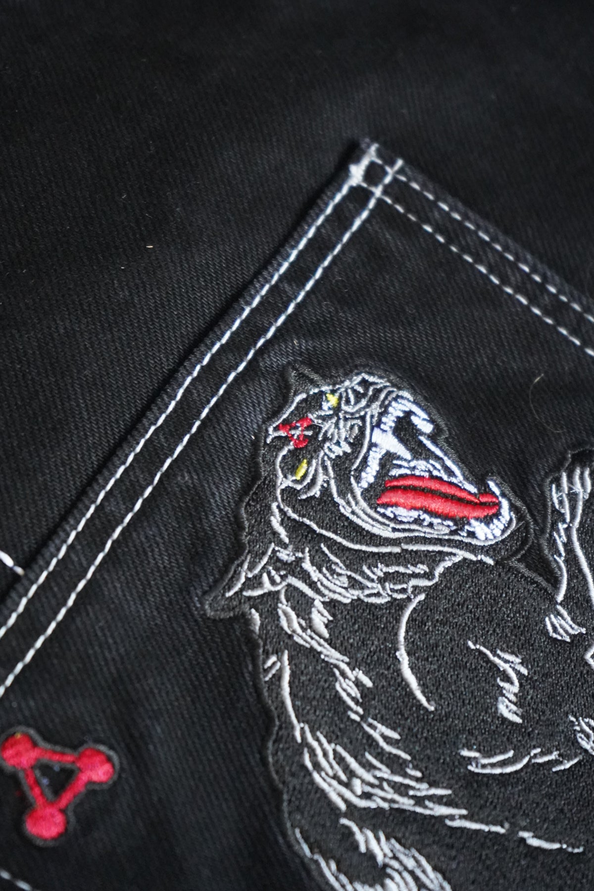 Close-up of the black Divine Dogs Pants back pocket with white contrast stitching, an embroidered snarling wolf-like dog patch and a small red triangle embroidery