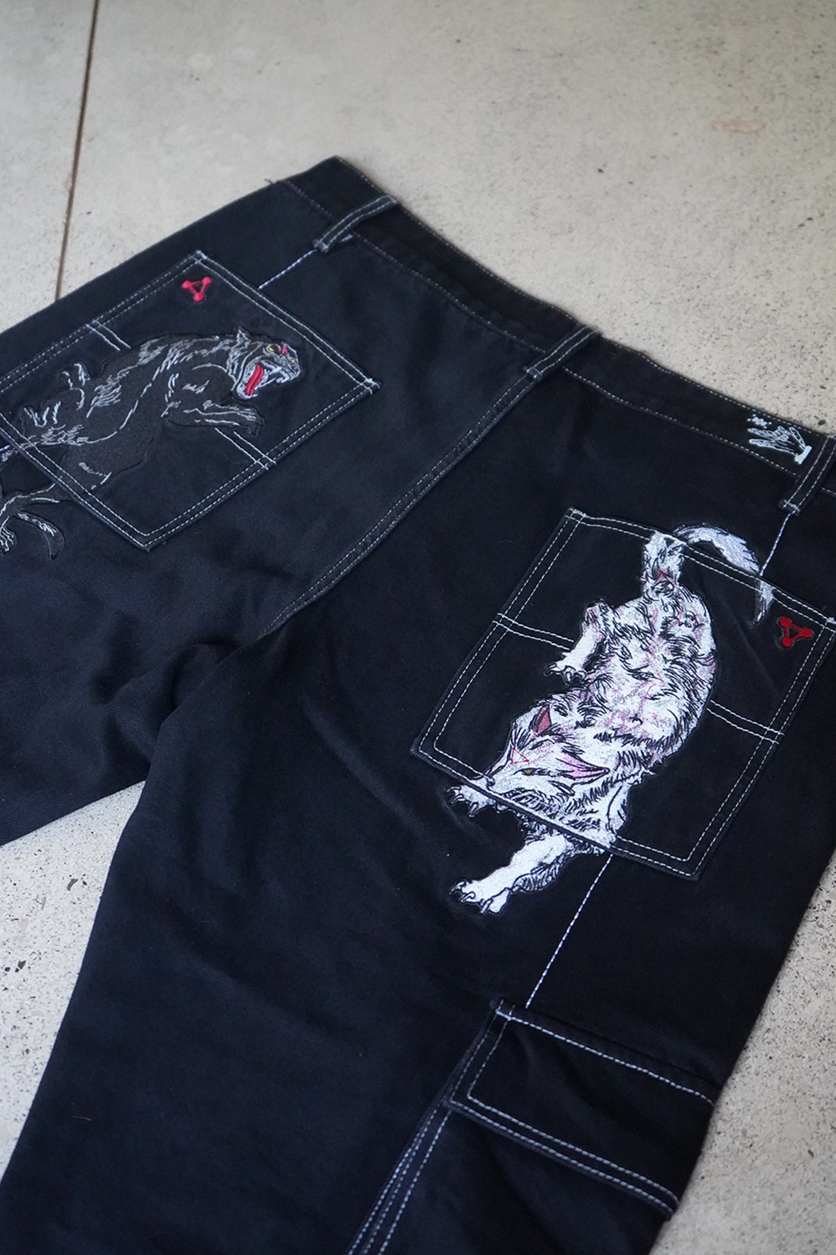 Close-up flat lay of the Divine Dogs Pants in black denim with white contrast stitching, showing embroidered wolf and panther graphics on the back pockets