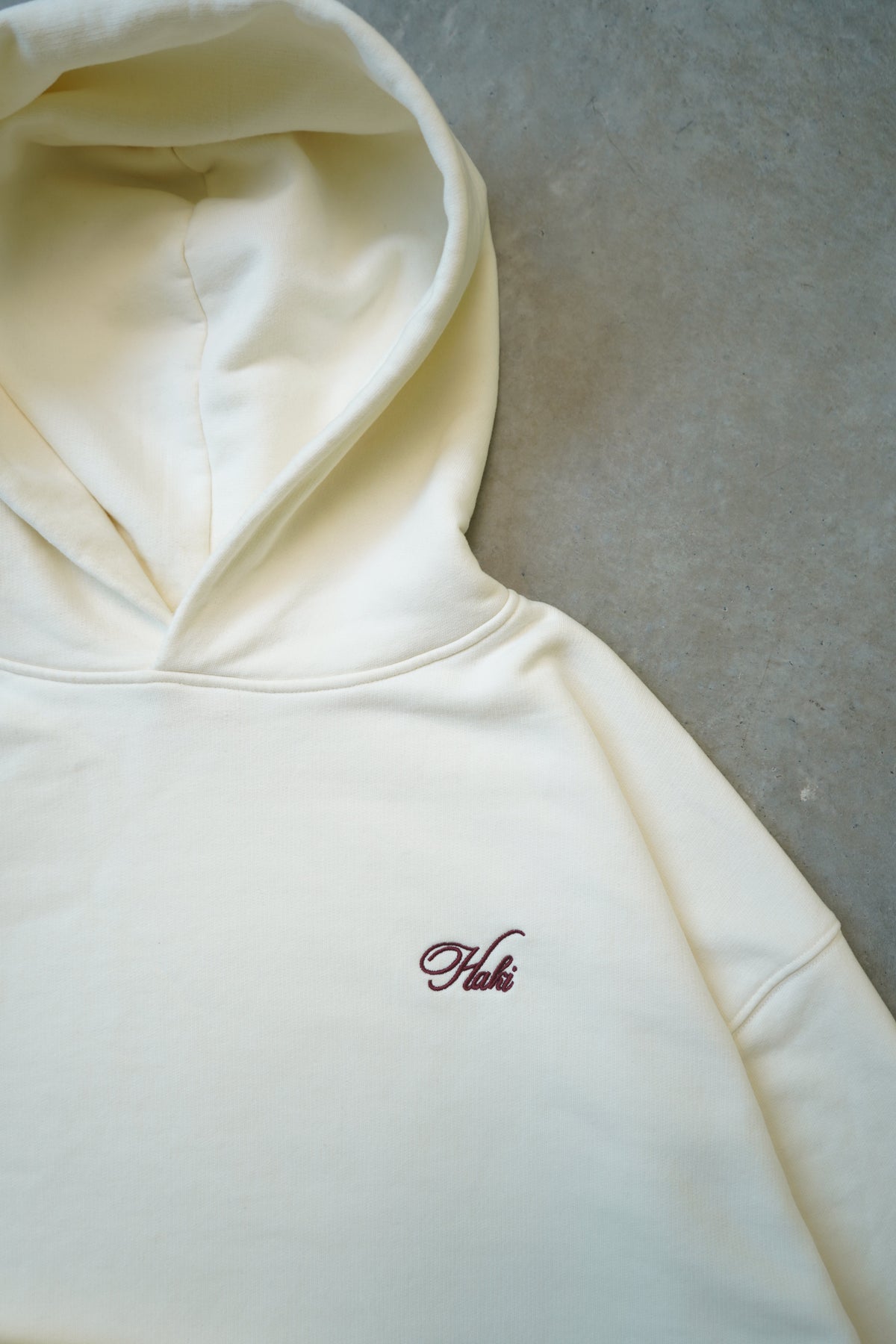 A cream-colored hoodie laid flat on a textured gray surface, featuring a small embroidered dark red script logo on the left chest.