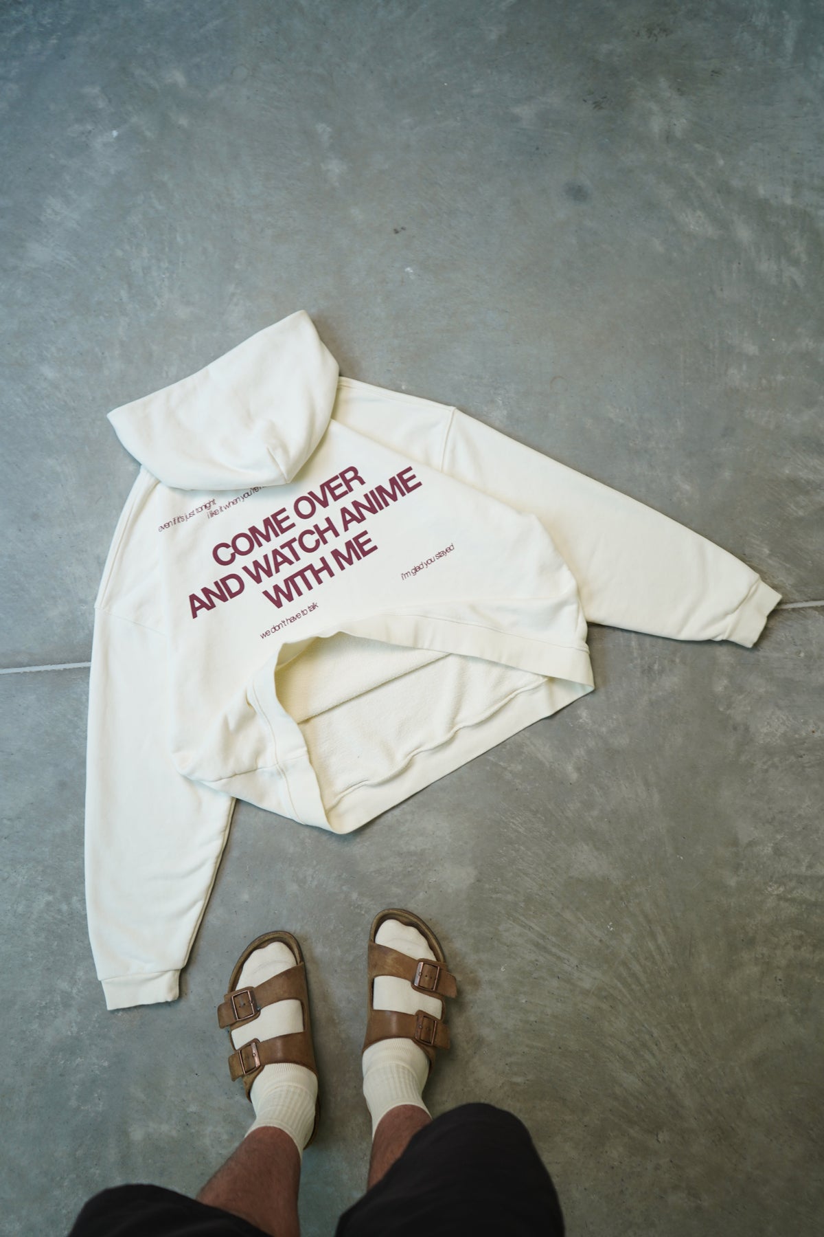 An off-white hoodie with red text reading 'COME OVER AND WATCH ANIME WITH ME' is laid out on a gray concrete floor, with a person's feet wearing brown slide sandals visible at the bottom.
