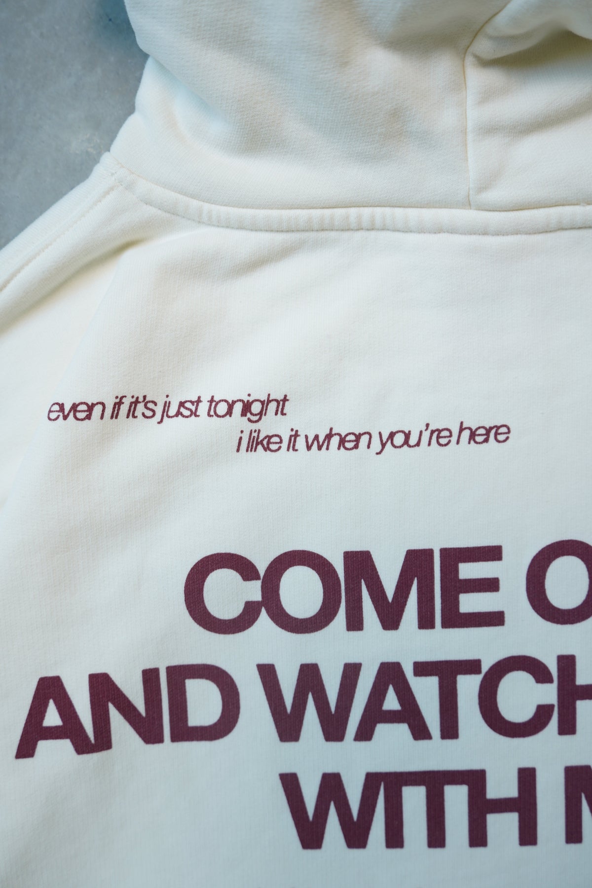Close-up of a cream-colored hoodie featuring dark burgundy text on the chest, including the phrase 'even if it's just tonight i like it when you're here' and large text reading 'COME ON AND WATCH WITH ME'.