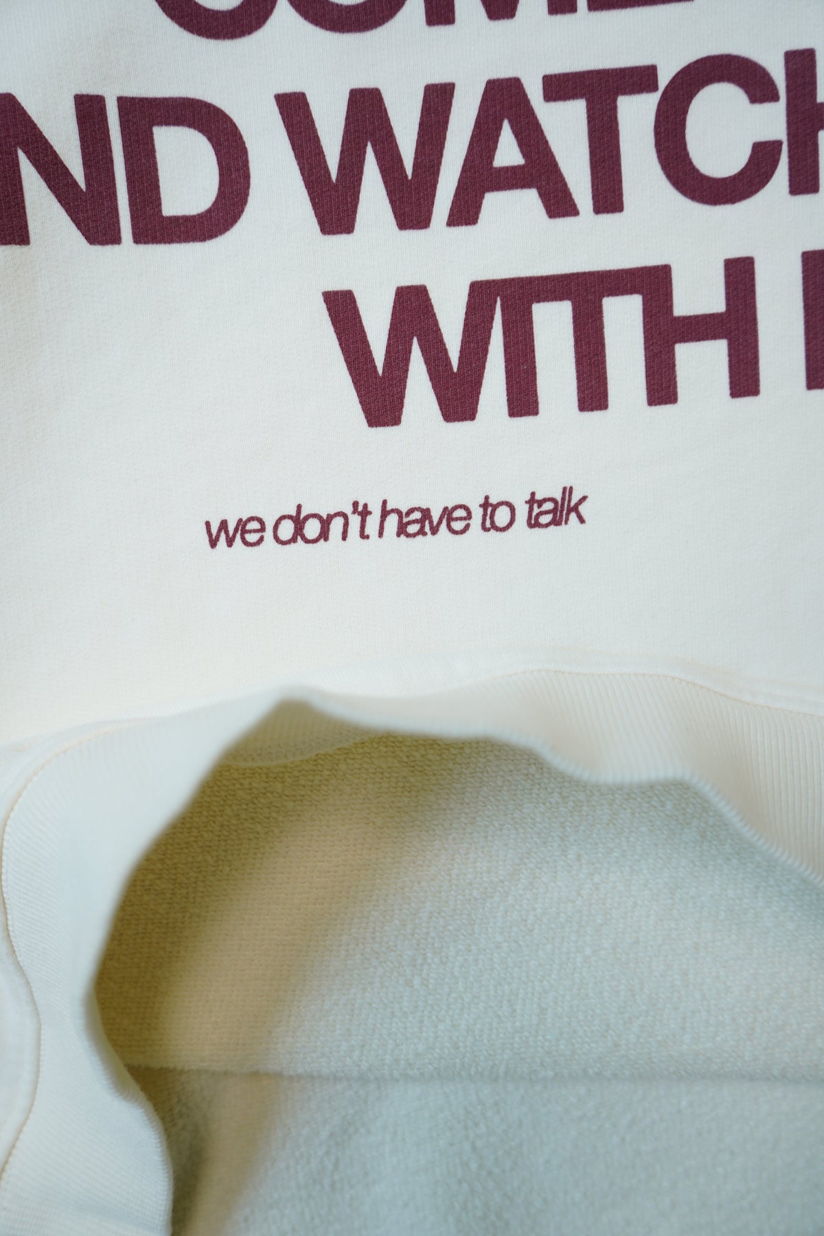 Close-up of a white sweatshirt with dark burgundy text reading 'COME AND WATCH WITH US' and the phrase 'we don't have to talk' printed below, with the hood opening shown.