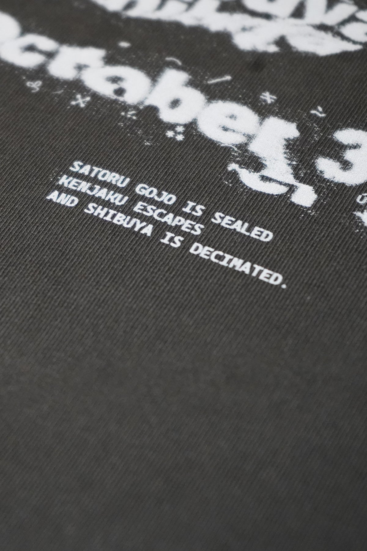 Close-up of the Shibuya Incident Tee in charcoal gray, with a distressed white graphic above small printed text reading Satoru Gojo is sealed, Kenjaku escapes and Shibuya is decimated