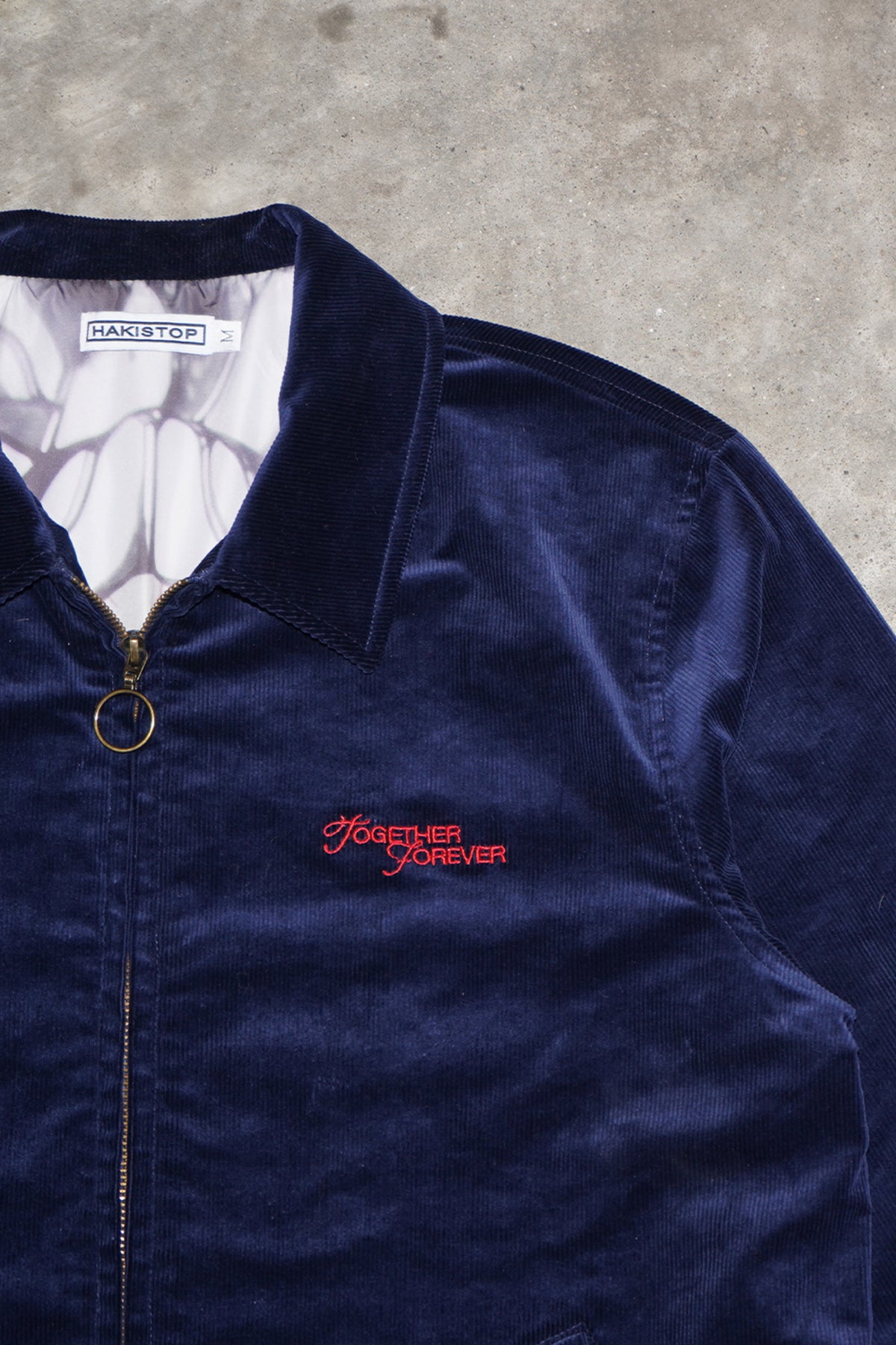 Close-up of the navy corduroy Queen of Curses Jacket showing collar, brass ring-pull zipper, red embroidered script on the chest and HAKISTOP label inside