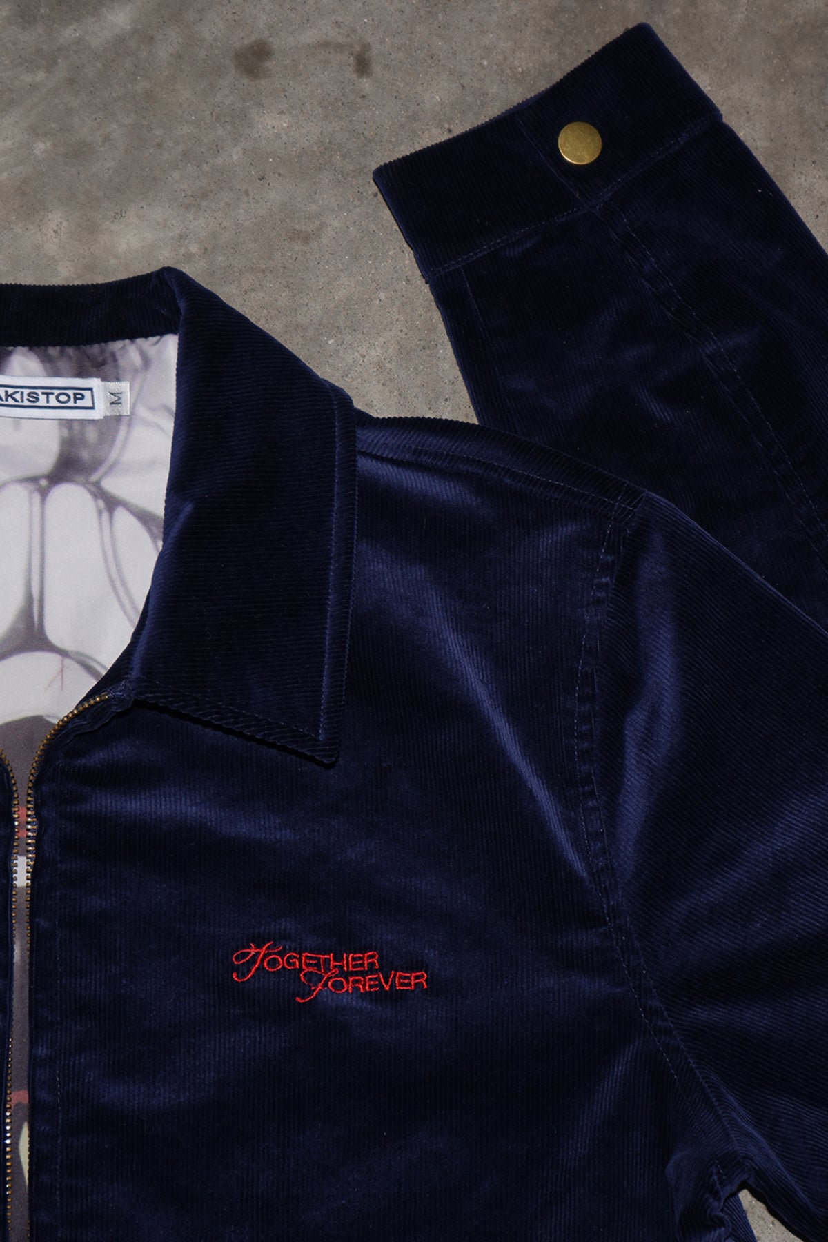 Flat lay close-up of the navy corduroy Queen of Curses Jacket showing collar, zipper, red embroidered "Together Forever" text, a gold snap cuff, and a graphic lining