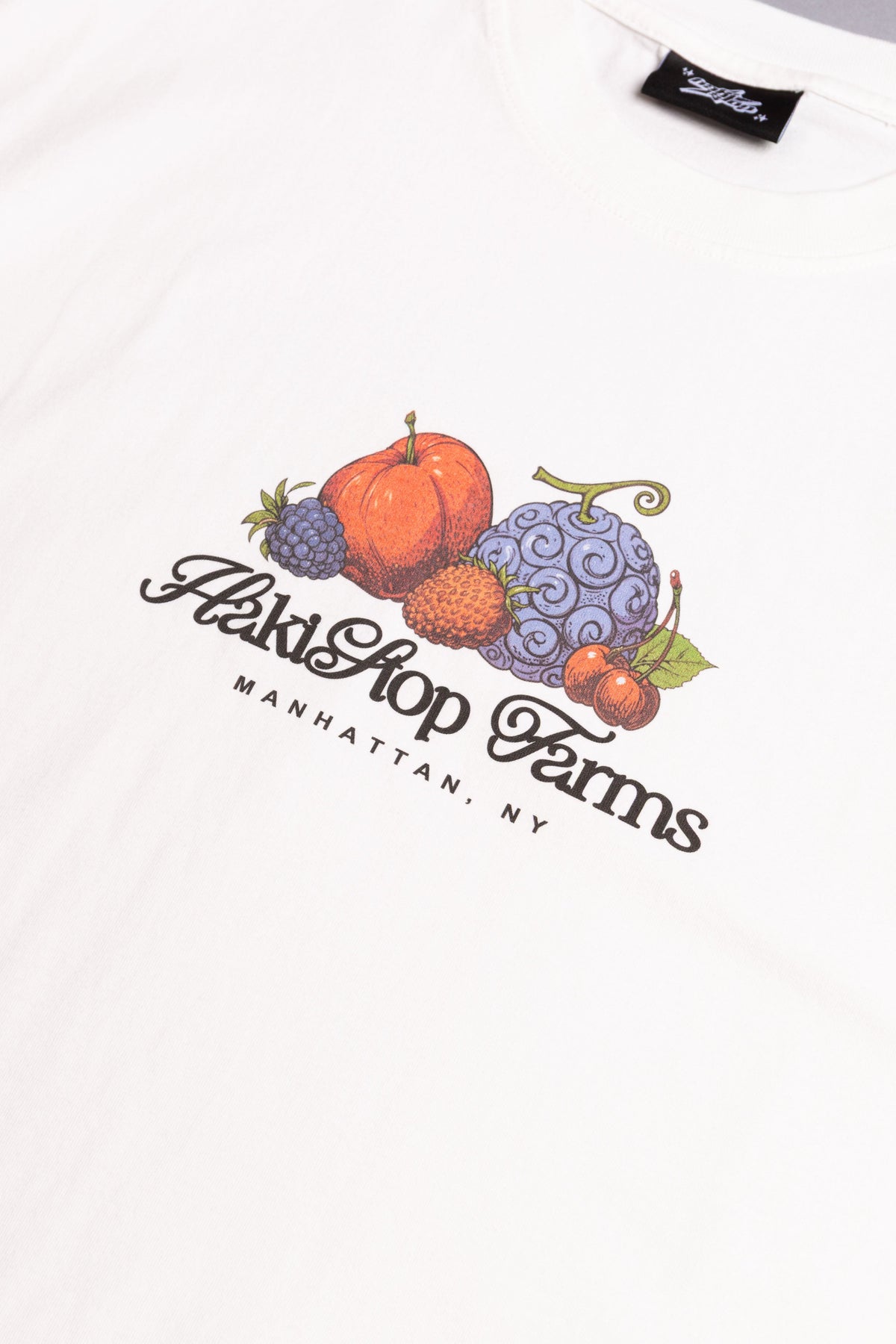Close-up of a white short-sleeve T-shirt with a colorful graphic print of various fruits and the text 'HakiStop Farms' and 'MANHATTAN, NY' on the chest.