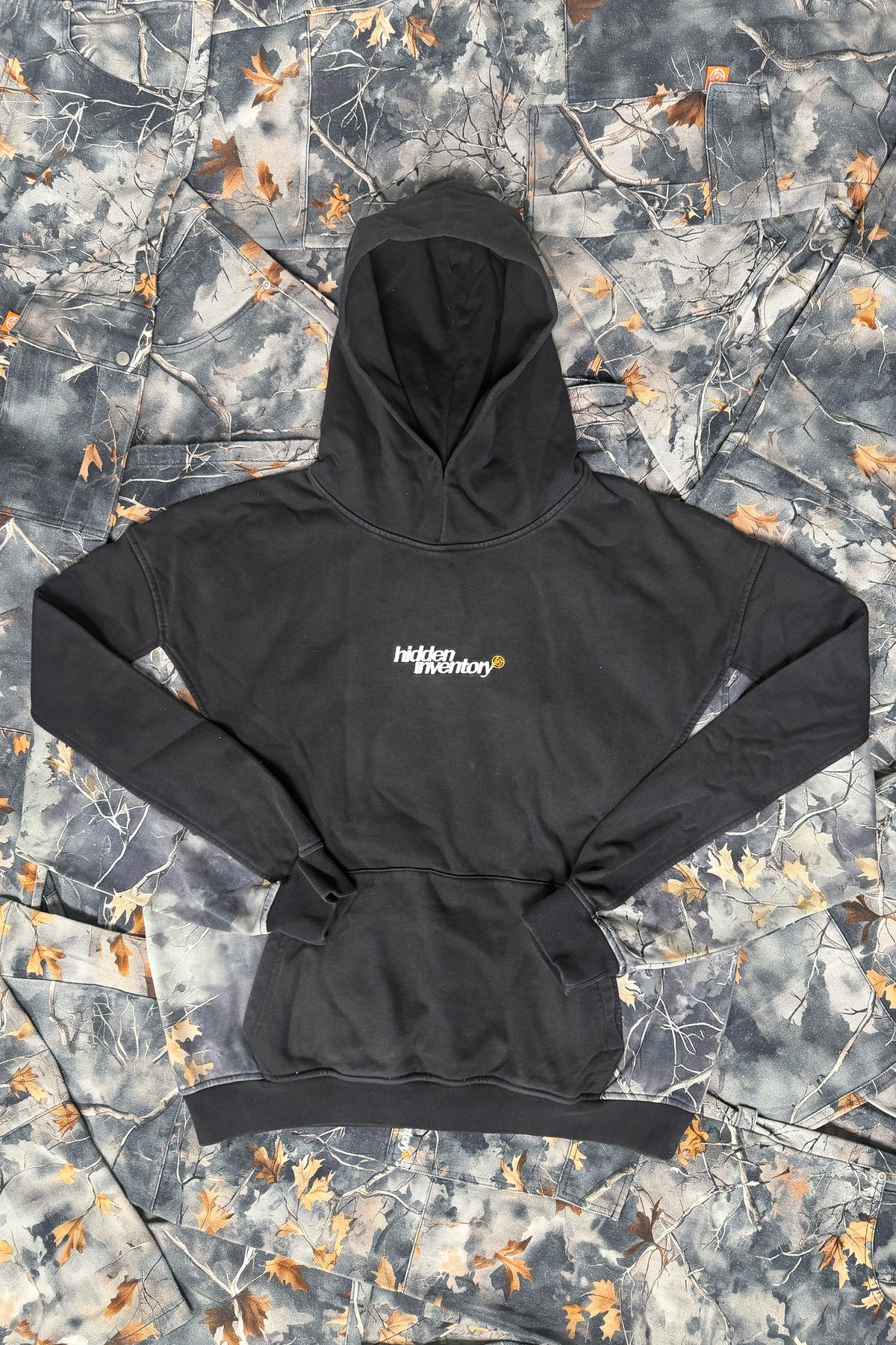 Flat lay front view of the black Hidden Inventory Hoodie with camouflage side panels, small embroidered chest text, and a kangaroo pocket, on camo fabric